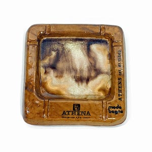 May include: A square ceramic ashtray with a glossy brown glaze and a central basin featuring a marbled cream and dark brown finish. The rim includes the text ATHENA CERAMICHE S.P.A. ITALY, ATHENS tet. 87.17.603, and the logo moda bagno.