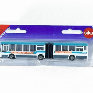 Op de afbeelding: Een speelgoed gelede busmodel in een doorzichtige plastic verpakking. De bus is wit, blauw en grijs, met de tekst "27" en "RATP" zichtbaar. De verpakking bevat de tekst "SIKU SUPER SERIE".