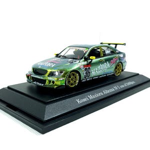 May include: A green and gold diecast model car with the number 36 on the side, displayed on a black base. The car has gold rims and a spoiler. The text "Kosei Maziora Altezza N-1" is printed on the base.