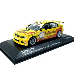 May include: A yellow BMW 320i model car with racing decals, including "Gaffelder" and "BERU" logos, sits on a black display stand. The model car has a spoiler and silver wheels. Text on the stand reads "BMW 320i" and "Deutsche Produktionswagen-Meisterschaft 2004 Champion Claudia Hürtgen."