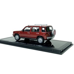 Könnte beinhalten: Ein weinrotes Modellauto eines Nissan Rasheen Type A 1997 auf einem schwarzen Präsentationsständer. Der Miniatur-SUV hat ein Reserverad hinten und einen silbernen Dachgepäckträger. Das Modell ist detailliert.