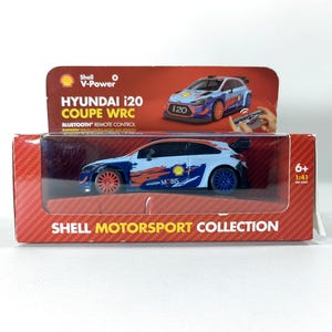 May include: A Hyundai i20 Coupe WRC model car in a red and clear plastic box. The car is white, blue, and orange with the text "MOBIS" and "Shell" on the side. The box reads "Shell Motorsport Collection".