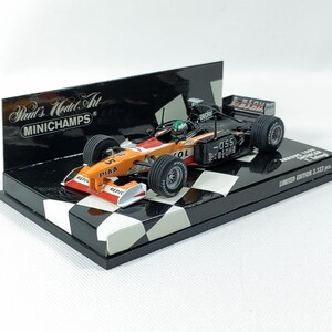 Può includere: Modello dettagliato di un'auto da corsa di Formula 1, prevalentemente arancione e nera, con loghi degli sponsor. Il modello è esposto su una base a scacchi in bianco e nero, con uno sfondo con il logo "MINICHAMPS". Edizione limitata.