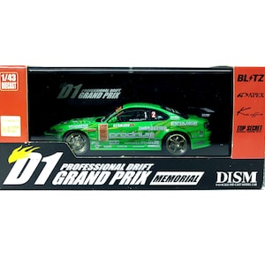 Op de afbeelding: Een groen en zwart modelauto in een displaydoos. De auto heeft sponsorlogo's en de tekst "D1 Grand Prix Memorial". De doos toont ook "1/43 Diecast" en "D1 Professional Drift Grand Prix".