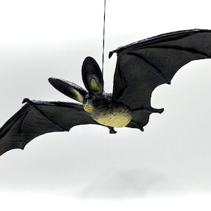 May include: A black bat decoration with outstretched wings, suspended against a white background. The bat has detailed wings and a yellow-toned body. The bat is a Halloween or spooky season decoration.