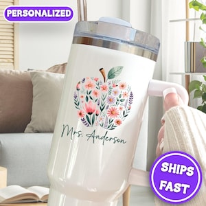 May include: White tumbler with a floral apple design and the text "Mrs. Anderson". The apple is filled with pink, purple, and white flowers. The tumbler has a handle and a straw. The word "PERSONALIZED" is in a purple banner.