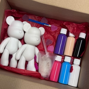 Può includere: Un kit per lavoretti creativi con due figurine di orso in ceramica bianca, sei bottiglie di vernice e un bicchiere di plastica con una sostanza bianca. I colori della vernice sono viola, rosa, blu, bianco e due tonalità di marrone. Il kit è confezionato in una scatola di cartone con carta velina rossa.