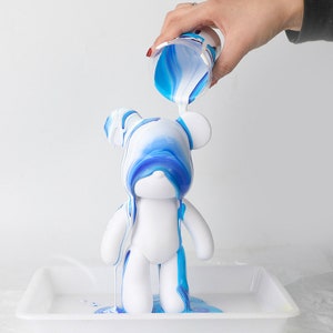 May include: A white ceramic bear figurine is being painted with blue and white paint. The paint is being poured from a cup and is dripping down the bear's body.