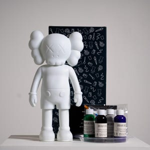 Può includere: Una figura di vinile bianca con un volto a forma di teschio e occhi a croce, accanto a una scatola nera con illustrazioni a tema artistico. La figura è accompagnata da vernici acriliche fluide in verde, nero, grigio e blu.