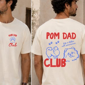 Pode incluir: Camiseta cor marfim com "POM DAD Club" impresso em vermelho e azul. A frente apresenta o texto e um desenho de pegada. A parte de trás exibe uma ilustração de Pomerânia e a frase "Life is better with Pomeranian."
