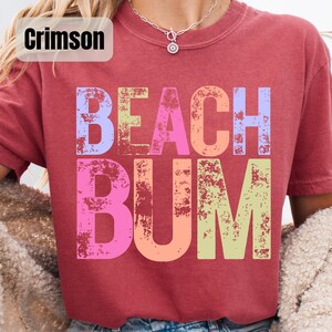 Op de afbeelding: Karmizijnrood T-shirt met de woorden "BEACH BUM" in grote, kleurrijke, vervaagde letters. De tekst is in tinten blauw, roze, oranje en groen. Het shirt heeft korte mouwen en een ronde hals.