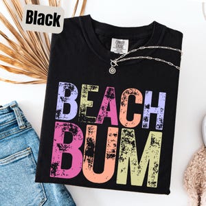 Op de afbeelding: Zwart T-shirt met de tekst "BEACH BUM" in kleurrijke, verweerde letters. Het shirt heeft een V-hals en een zilveren ketting. Het shirt is opgevouwen op een witte ondergrond naast een spijkerbroek en een beige trui.
