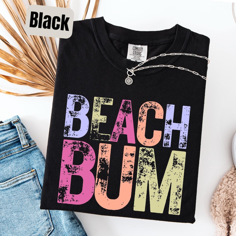 Op de afbeelding: Zwart T-shirt met de tekst "BEACH BUM" in kleurrijke, verweerde letters. Het shirt heeft een V-hals en een zilveren ketting. Het shirt is opgevouwen op een witte ondergrond naast een spijkerbroek en een beige trui.