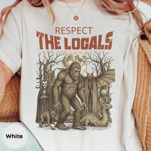 May include: White t-shirt with the text "RESPECT THE LOCALS" above a graphic of various cryptids, including Bigfoot, a vampire, and a winged creature, under a full moon.