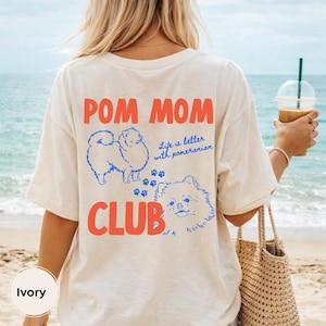 Pode incluir: Camiseta cor marfim com as palavras "POM MOM CLUB" em vermelho e azul. A camisa apresenta ilustrações azuis de cães da raça Pomerânia e a frase "Life is better with pomeranian."