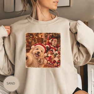 May include: A sand-coloured crewneck sweatshirt featuring a graphic print of a golden-coloured dog wearing a red Santa hat and holding a mug, with Santa Claus sitting in the background amidst Christmas decorations and wrapped gifts.
