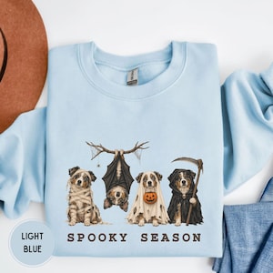 Pode incluir: Uma camisola de gola redonda azul-clara com um gráfico de quatro cães com fatos de Halloween. Um cão é uma múmia, um está pendurado como um morcego, um usa um lençol de fantasma com um balde de abóbora e um é a morte. Texto: SPOOKY SEASON.