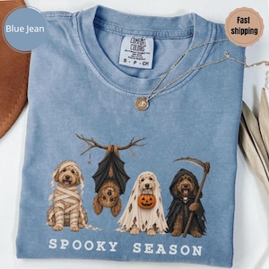 Może przedstawiać: Niebieska koszulka z grafiką przedstawiającą cztery psy w kostiumach na Halloween: mumię, nietoperza wiszącego na gałęzi, ducha trzymającego dynię oraz ponurego żniwiarza z kosą. Poniżej psów znajduje się nadruk z napisem "SPOOKY SEASON".