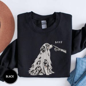 Könnte beinhalten: Ein schwarzes Sweatshirt mit Rundhalsausschnitt und einer Grafik eines Australian Shepherd, der ein weißes Geisterlaken mit Löchern trägt. Ein Skelettfinger berührt die Nase des Hundes, darüber steht das Wort BOOP in weißer Schrift.