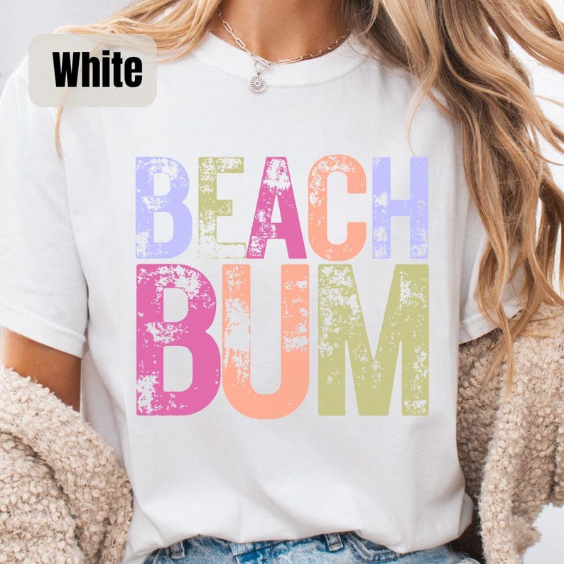 Op de afbeelding: Wit T-shirt met de woorden "BEACH BUM" in grote, kleurrijke letters. De tekst heeft een verweerde, getextureerde uitstraling, waarbij elk woord een andere pastelkleur heeft. Een casual kledingstuk met een zomerthema.