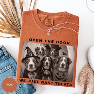 Può includere: Una t-shirt color arancio bruciato con una grafica in bianco e nero di quattro cani in costume di Halloween. Il testo recita OPEN THE DOOR WE JUST WANT TREATS. La maglietta presenta un girocollo e un'etichetta Comfort Colors sul colletto.