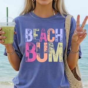 Op de afbeelding: Een blauw T-shirt met de tekst "BEACH BUM" in kleurrijke, vervaagde letters. Het shirt heeft een relaxte pasvorm. De afbeelding toont ook een persoon die een groen drankje met een rietje vasthoudt.