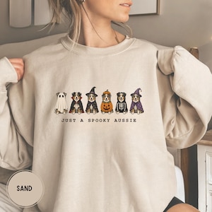 Può includere: Una felpa girocollo color sabbia con una grafica di sei cani pastore australiano in vari costumi di Halloween, tra cui un fantasma, un vampiro, una strega, una zucca, uno scheletro e un mago. Il testo sotto i cani recita JUST A SPOOKY AUSSIE.
