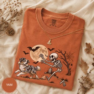 Peut inclure: Un t-shirt orange brûlé avec un motif représentant un squelette tendant la main vers un berger australien tenant un os. La scène inclut une pleine lune, des chauves-souris, un arbre dénudé avec une toile d'araignée et une pierre tombale. Un collier chapeau de sorcière doré repose sur le t-shirt.