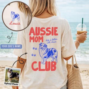 Puede incluir: Una camiseta de color crema con un gráfico en la espalda que incluye el texto rojo "AUSSIE MOM CLUB" y una ilustración azul de un perro pastor australiano. El texto "Life is better with Louie" está impreso en azul junto a huellas azules.