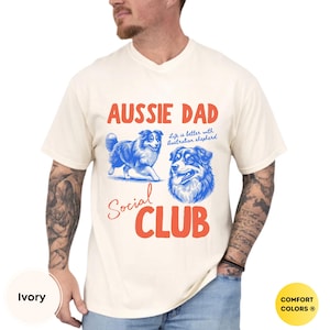 May include: Cream-coloured t-shirt with the text "AUSSIE DAD Social CLUB" in red and blue. The shirt features illustrations of an Australian Shepherd dog. The phrase "Life is better with Australian shepherd" is also printed on the shirt.