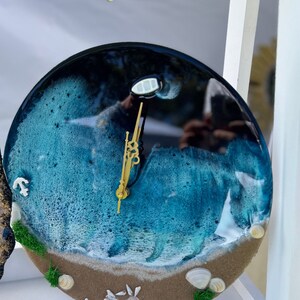 Op de afbeelding: Een ronde harsklok met een strandscene. De wijzerplaat van de klok is blauw en wit, met een zandstrand aan de onderkant. De wijzers van de klok zijn goudkleurig. De klok hangt aan een witte muur.