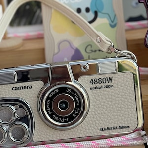 Könnte beinhalten: Ein silbernes und weißes Handy-Etui in Form einer Kamera mit einem Riemen. Das Etui hat ein Objektiv mit dem Text "4880W optical 200m" und "13.8-15.5 124-450mm".
