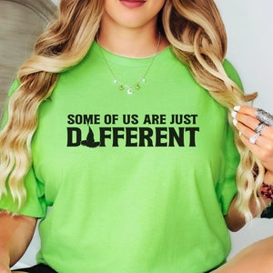 May include: A lime green t-shirt with the text "SOME OF US ARE JUST DIFFERENT" in black. The shirt features a witch hat graphic. The person is wearing black shorts and sitting on a white bed.