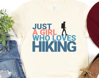 May include: A beige t-shirt with the text "Just a girl who loves hiking" in blue, pink, and white. A silhouette of a person hiking is to the right of the text.