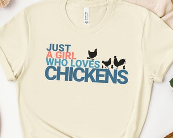 May include: A beige t-shirt with the text "Just a girl who loves chickens" in blue and pink lettering. There are black silhouettes of chickens below the text.