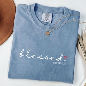 May include: A blue Comfort Colors t-shirt with the word "blessed" in white script, a small red heart, and the text "Jerimiah 17:7". A gold necklace with a pendant rests on the shirt. The shirt is folded.