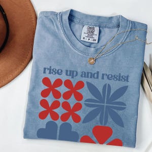 May include: A blue Comfort Colors t-shirt with the words "rise up and resist" and floral graphics in red and blue. A gold necklace with a pendant is draped across the shirt.