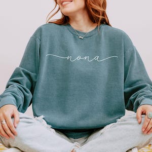 May include: Teal crewneck sweatshirt with the word "nona" in a white script font. The sweatshirt is a solid teal color and has long sleeves. The word "nona" is written in a cursive style.
