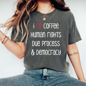 May include: Dark grey t-shirt with the text "I ❤️ COFFEE HUMAN RIGHTS DUE PROCESS & DEMOCRACY" in white. The heart symbol is red. The shirt is a casual style.