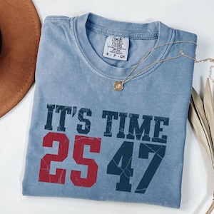 May include: A blue Comfort Colors t-shirt with the text "IT'S TIME" in dark blue and the numbers "25" in red and "47" in dark blue. A gold necklace with a pendant is on the shirt.