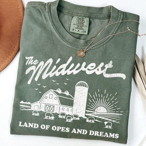 Op de afbeelding: Een olijfgroen T-shirt met korte mouwen en een witte afbeelding van een schuur, silo en zon. De tekst luidt The Midwest Land of Opes and Dreams. Een goudkleurige ketting met een kleine ronde hanger rust op de halslijn van het shirt.
