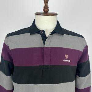 May include: A long-sleeved rugby shirt with horizontal stripes in black, gray, and purple. The shirt has a black collar and a small Guinness logo on the chest.