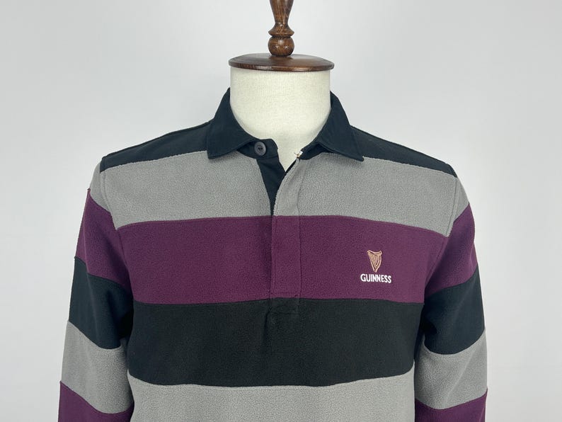 May include: A long-sleeved rugby shirt with horizontal stripes in black, gray, and purple. The shirt has a black collar and a small Guinness logo on the chest.