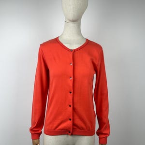 May include: A vibrant orange cardigan with a round neckline and long sleeves. The button-down sweater features black buttons and is displayed on a mannequin. The garment is made of a soft material.