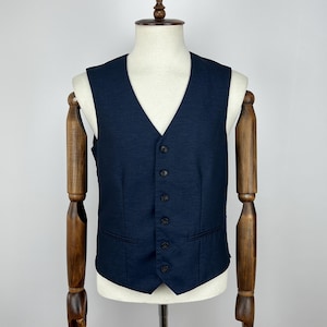 May include: A navy blue vest with a V-neck and six dark buttons. The vest has two front pockets and is displayed on a mannequin with wooden arms. The background is plain white.