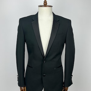 May include: A black blazer with a satin lapel and two-button closure. The jacket is displayed on a mannequin, showcasing its tailored fit and design. The sleeves have decorative buttons.