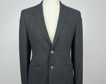 Könnte beinhalten: Ein dunkelgrauer Blazer auf einer Schaufensterpuppe. Der Blazer hat einen Zwei-Knopf-Verschluss, fallendes Revers und zwei Vordertaschen. Der Stoff hat ein subtiles strukturiertes Muster. Die Schaufensterpuppe steht vor einem weißen Hintergrund.