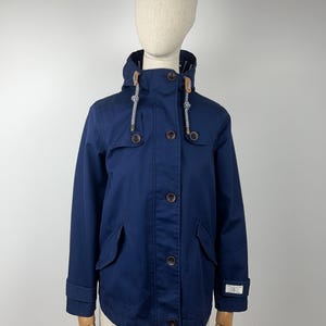 Puede incluir: Chaqueta azul marino con capucha, botones de madera y dos bolsillos delanteros. La capucha tiene cordones blancos y azules. La chaqueta se muestra en un maniquí.
