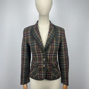 May include: A fitted blazer featuring a dark grey, red, and blue plaid pattern. The garment includes a notched lapel, three front buttons, and two flap pockets at the waist. The edges are finished with dark grey piping along the lapels and pockets.