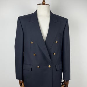 Puede incluir: Un blazer azul marino de doble botonadura con botones dorados. El blazer tiene una solapa de muesca clásica, dos bolsillos delanteros y se exhibe en un maniquí. La chaqueta es de corte entallado.